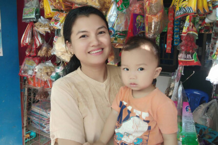 une bénéficiaire de Sont Oo Tehtwin pose devant sa boutique, avec son bébé dans les bras